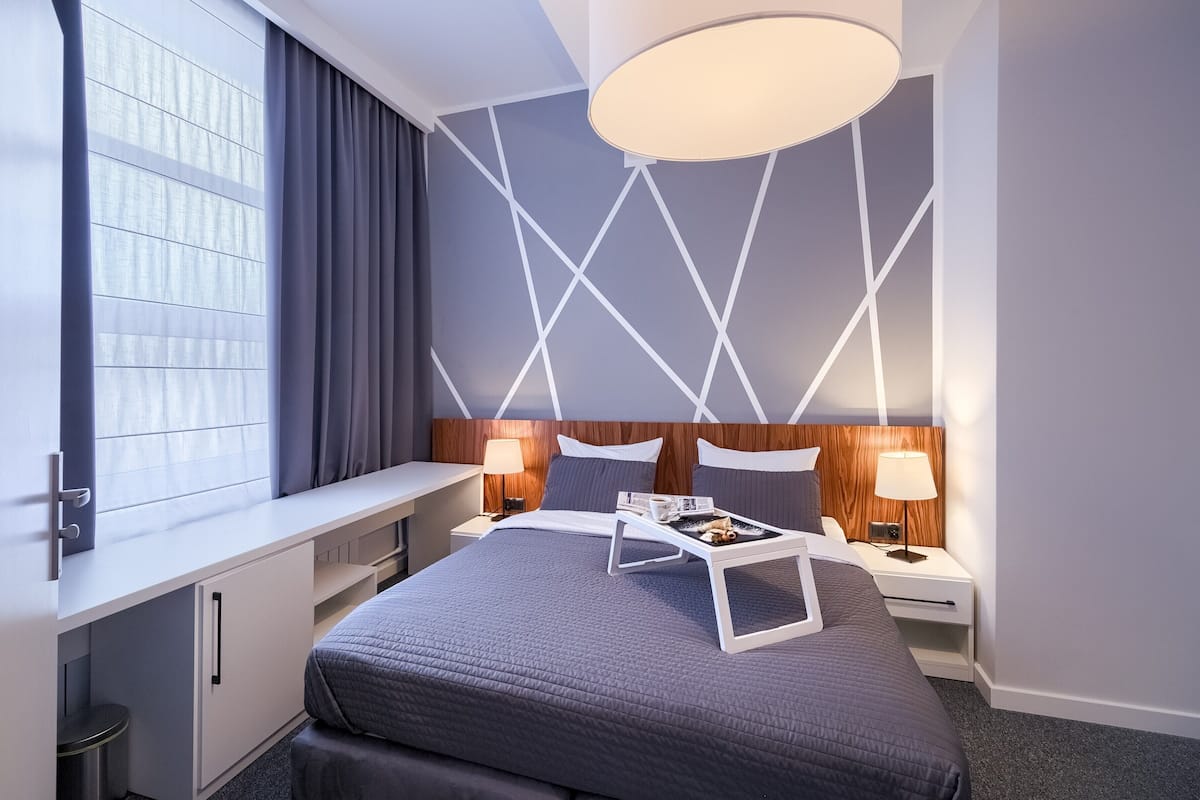 A modern hotel room with a large bed, bedside tables, a desk, and a window with curtains.