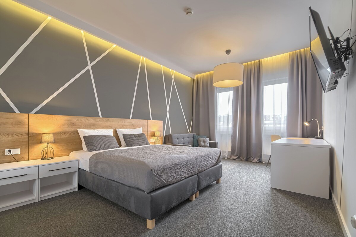 Una habitación de hotel moderna con una cama grande, mesitas de noche, un escritorio, un televisor y una ventana con cortinas.