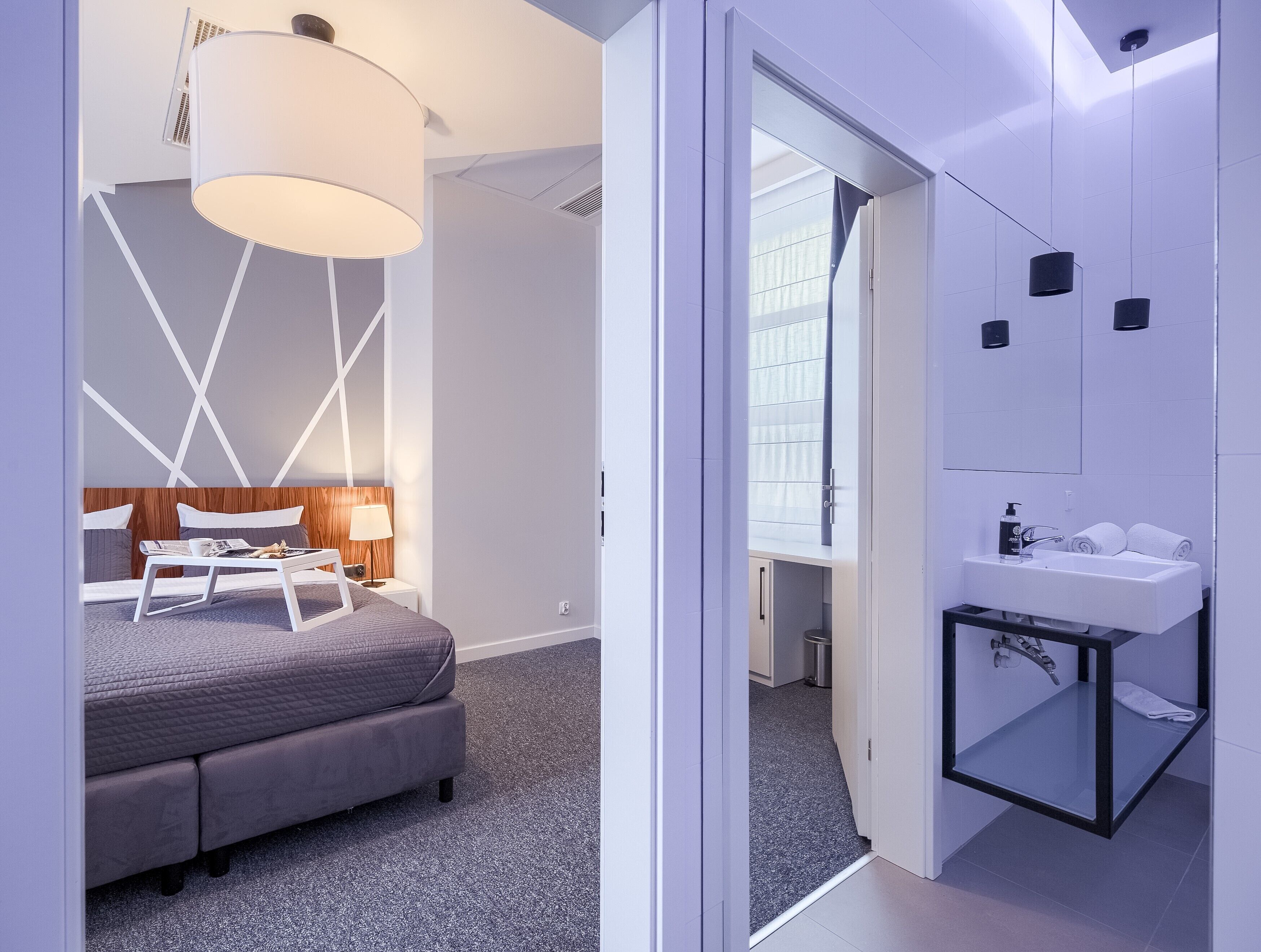 Quarto moderno com cama, mesa de cabeceira, luminária e vista para o banheiro.