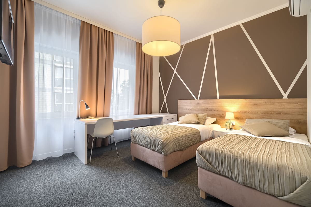 A hotel room with a desk, two beds, a TV, and a large window with curtains.