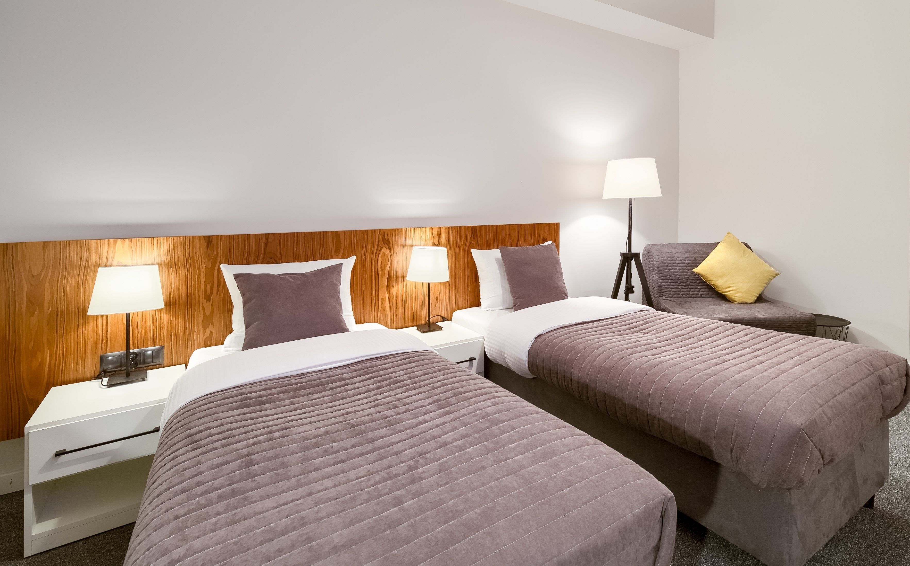 A hotel room with two beds, a wooden headboard, bedside lamps, a nightstand, and a chair with a cushion.