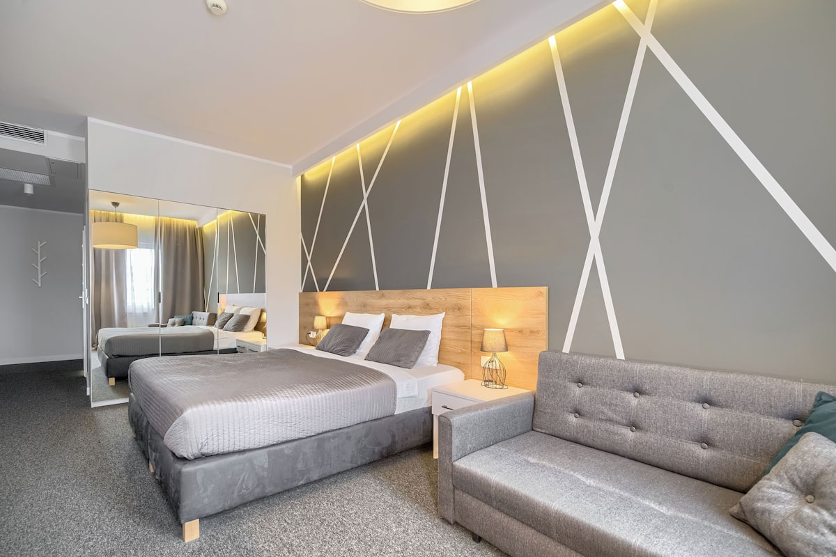 A modern hotel room with two beds, a sofa, a large mirror, and a geometric wall design.