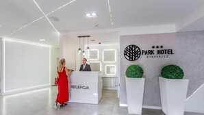 A modern hotel reception area with a reception desk labeled "RECEPTION," a man standing behind it, and a wall with the hotel name "PARK HOTEL BYDGOSZCZ" and a logo.