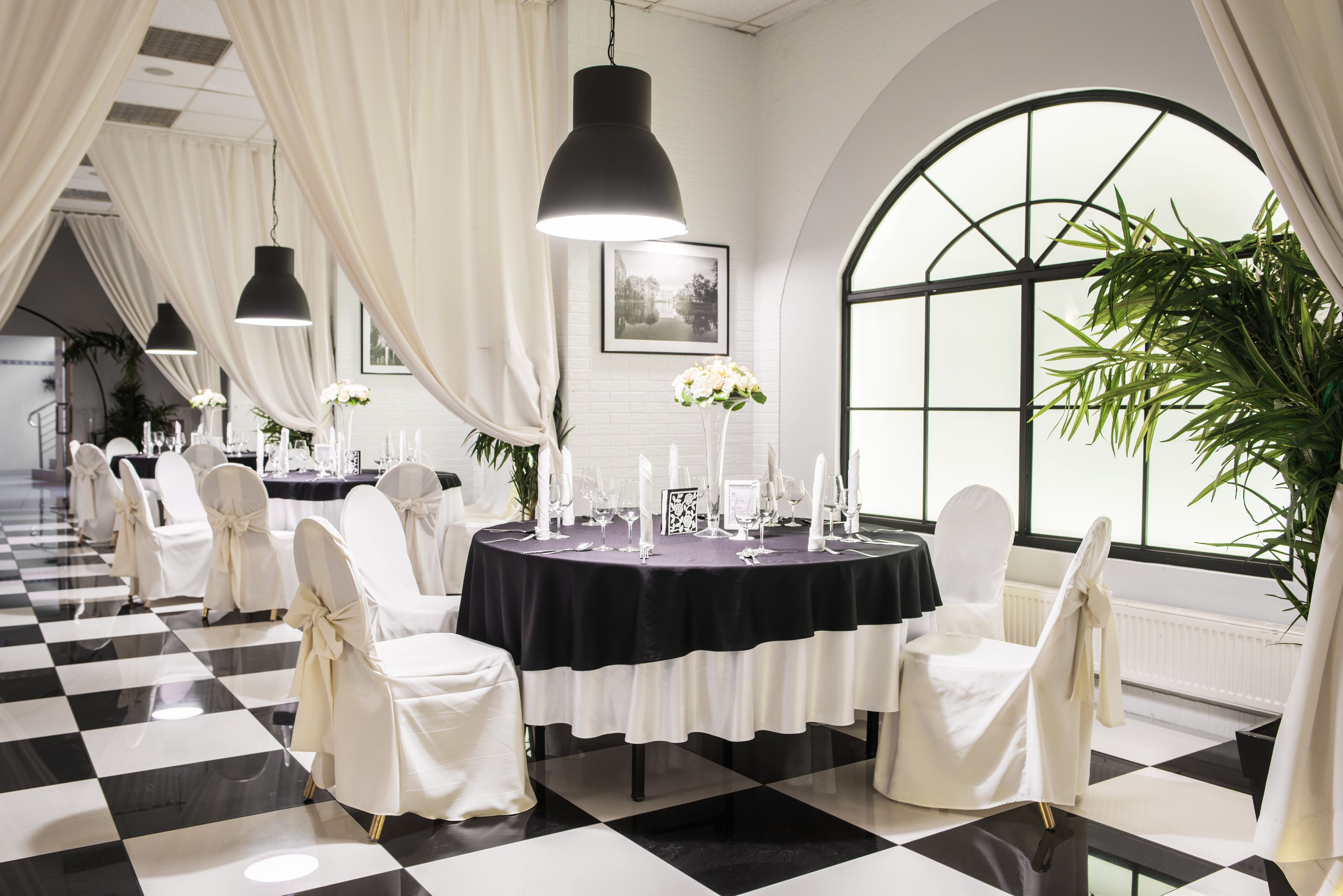A formal dining area with round tables, white chairs, black and white checkered flooring, and large windows with curtains.