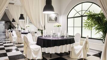 A formal dining area with round tables, white chairs, black and white checkered flooring, and large windows with curtains.