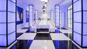 Una larga mesa de comedor con manteles blancos y sillas, ubicada en una habitación moderna con iluminación azul y ventanas amplias.