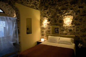A bedroom with a stone wall, a bed with white linens, a nightstand with a lamp, and a window with a sheer curtain.