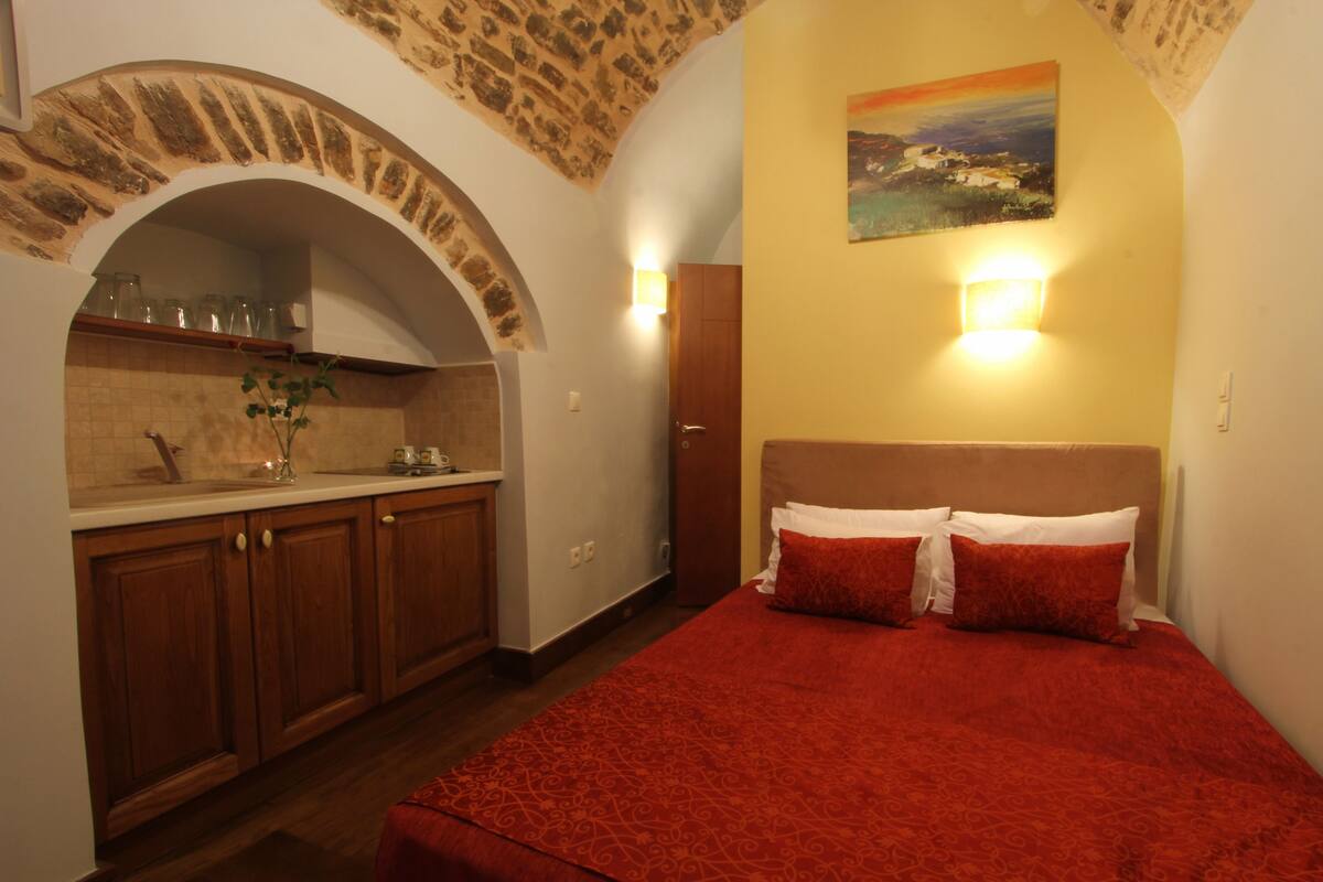 A bedroom with a red bedspread, a painting on the wall, and a kitchen area with wooden cabinets and a stone archway.
