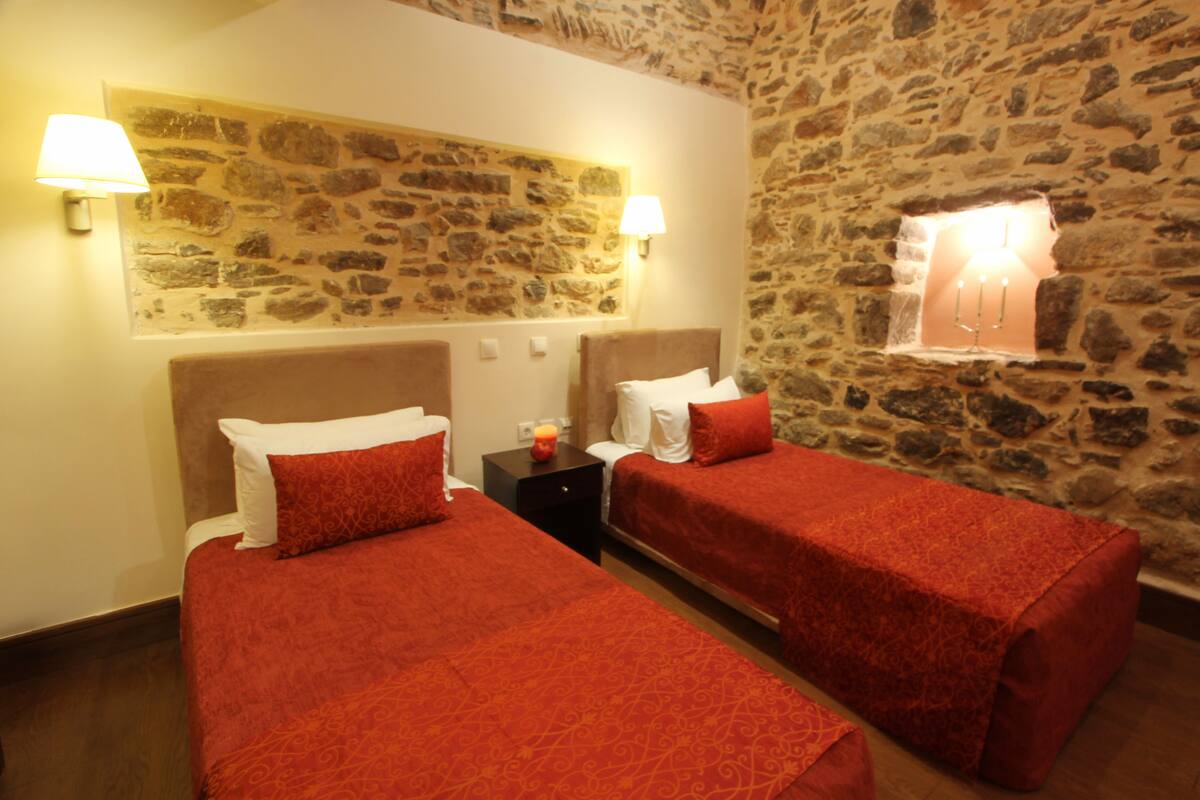 A room with two beds, each with a red bedspread and white pillows. There are two wall-mounted lamps on either side of the beds. The walls are made of stone, and there is a small window with a lamp inside.