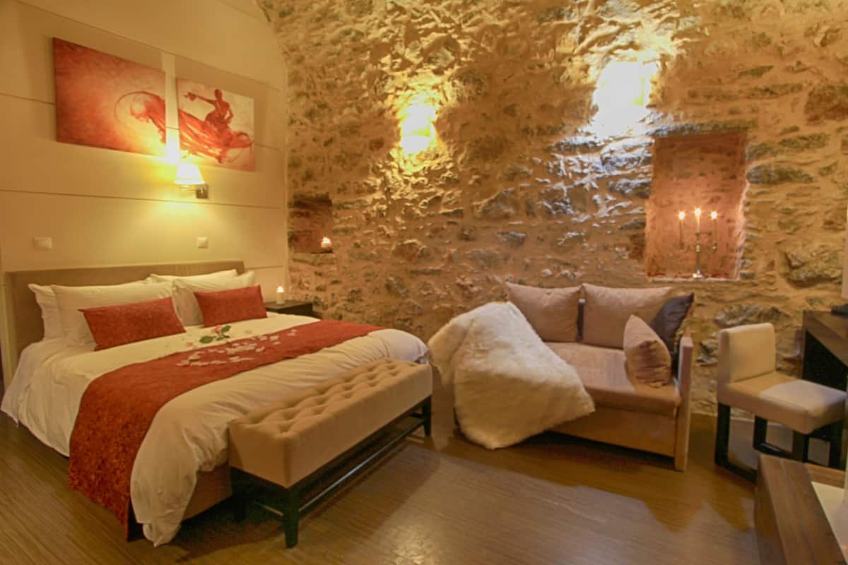 A bedroom with a stone wall, a bed with white bedding and red accents, a white armchair, and a wooden nightstand.