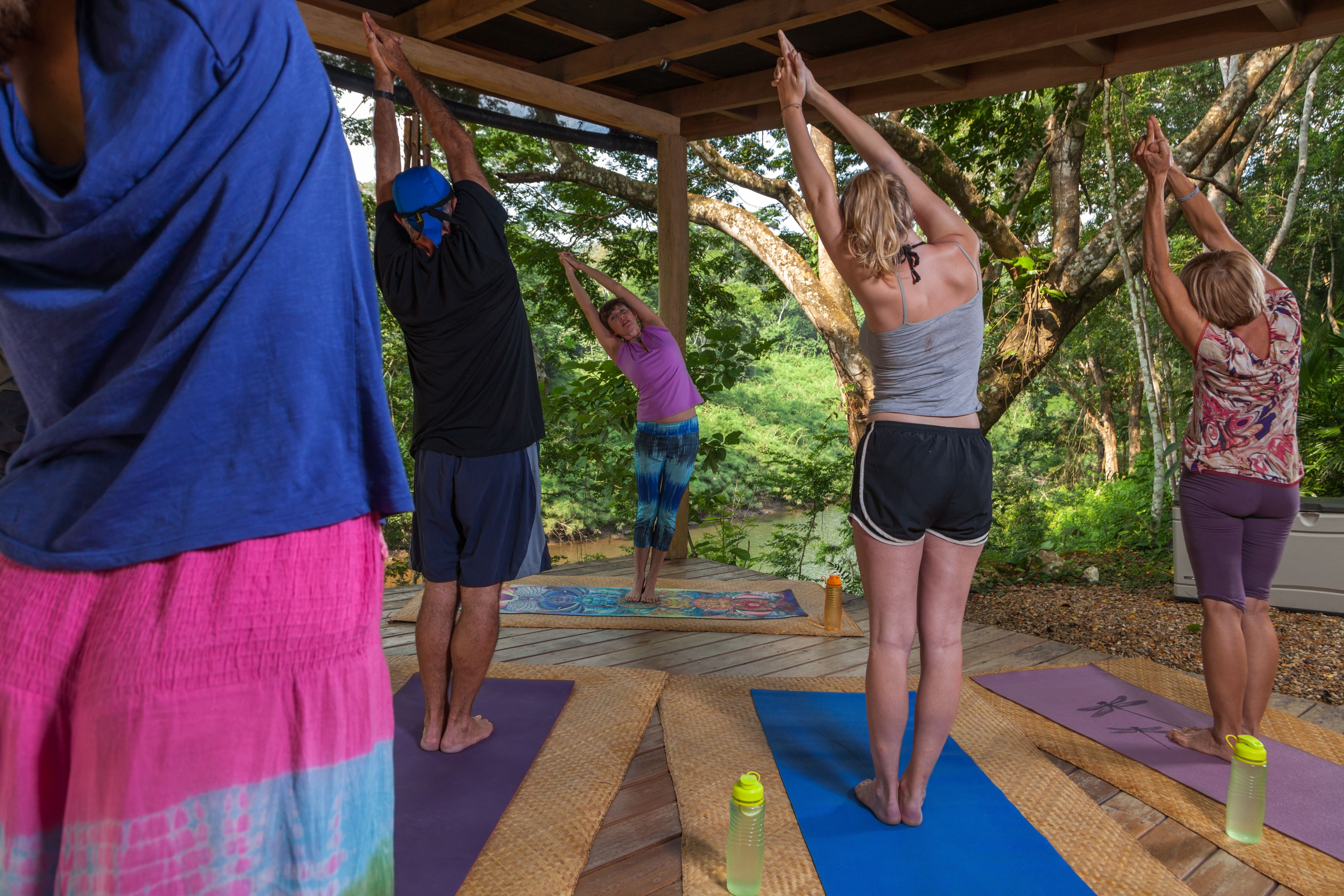 Un groupe de personnes pratiquant le yoga en plein air, sur des tapis, sous une structure en bois entourée d’arbres.