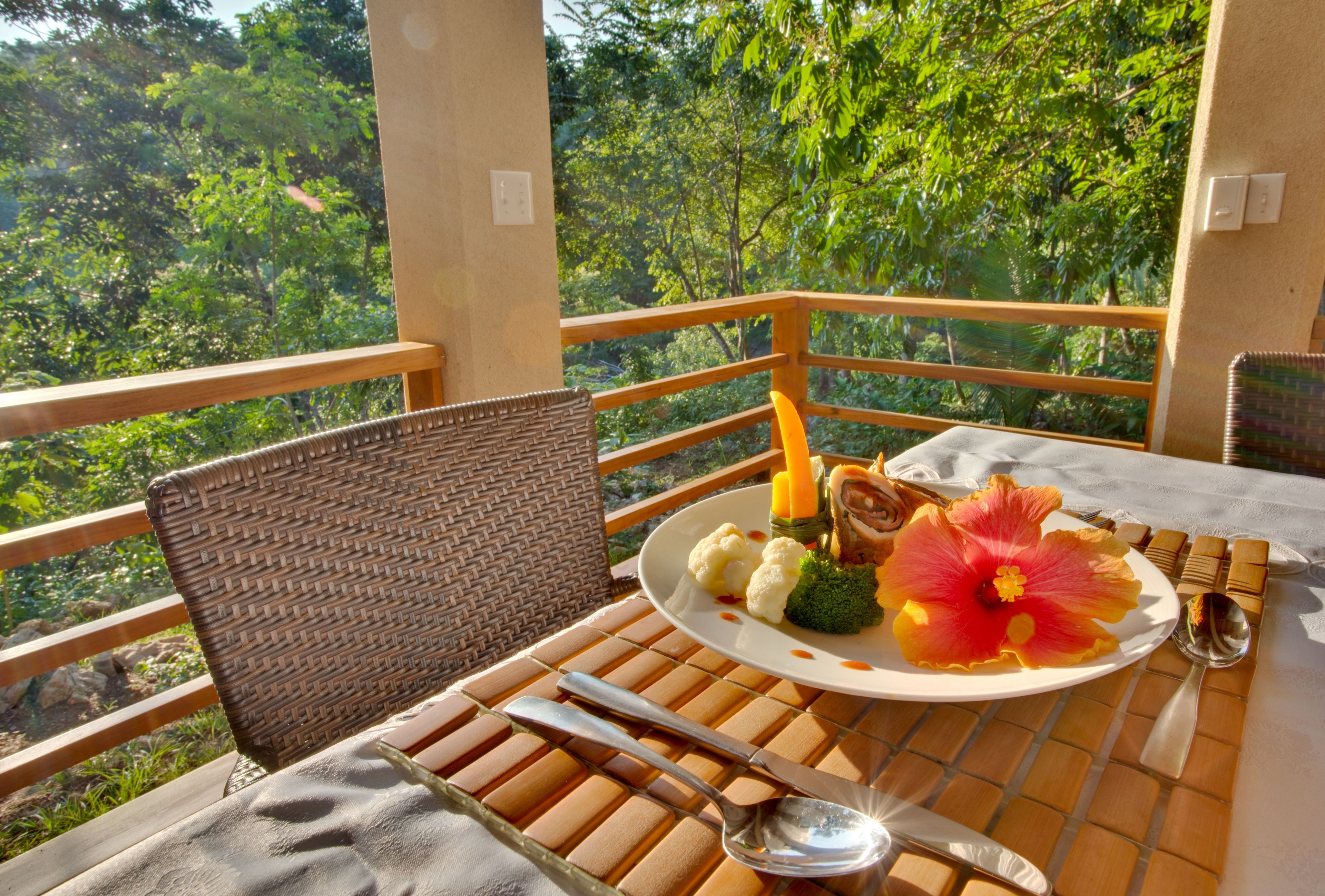 A dining table with a plate of food, cutlery, and a chair on a balcony with a view of trees.