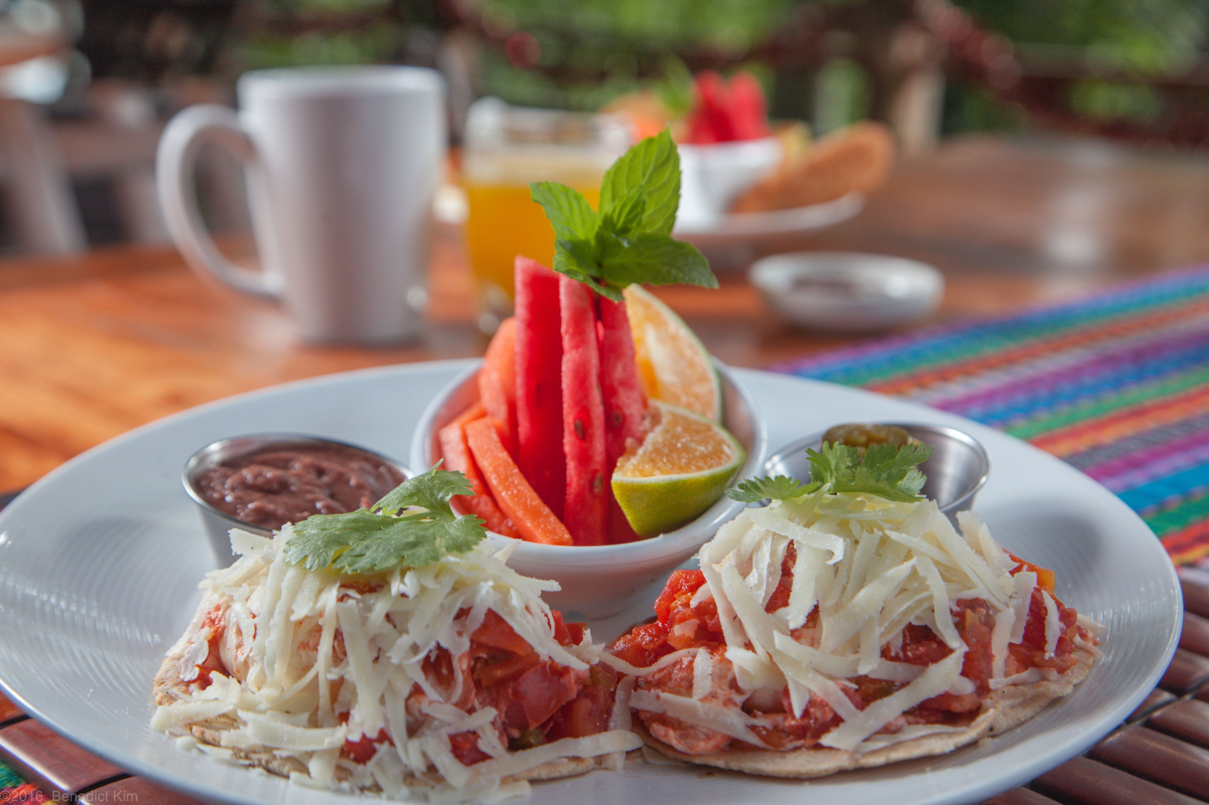 A plate with two tacos, a slice of watermelon, a lime, and a small bowl of beans.