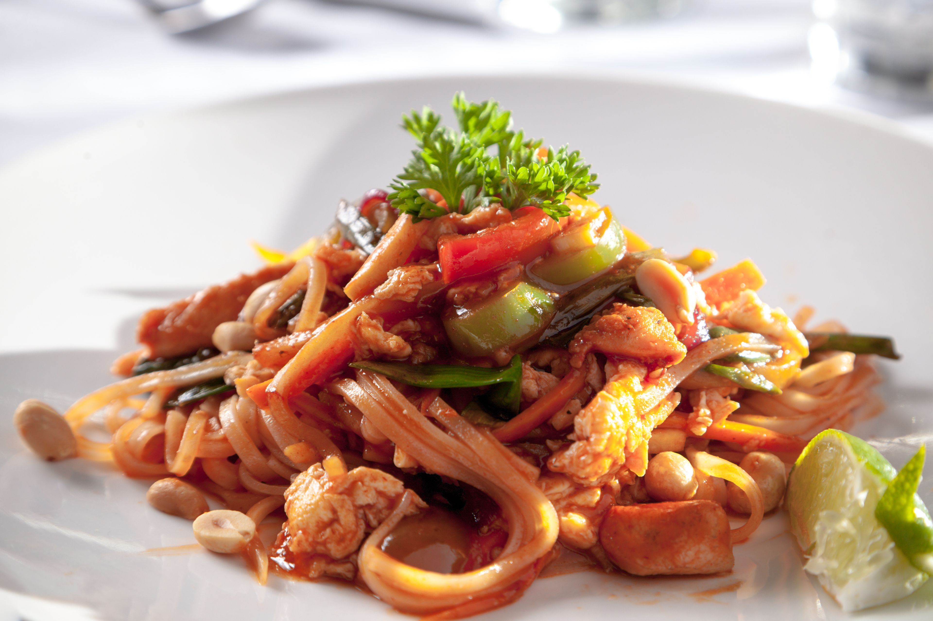 A plate of stir-fried noodles with vegetables and peanuts, garnished with parsley and a lime wedge.