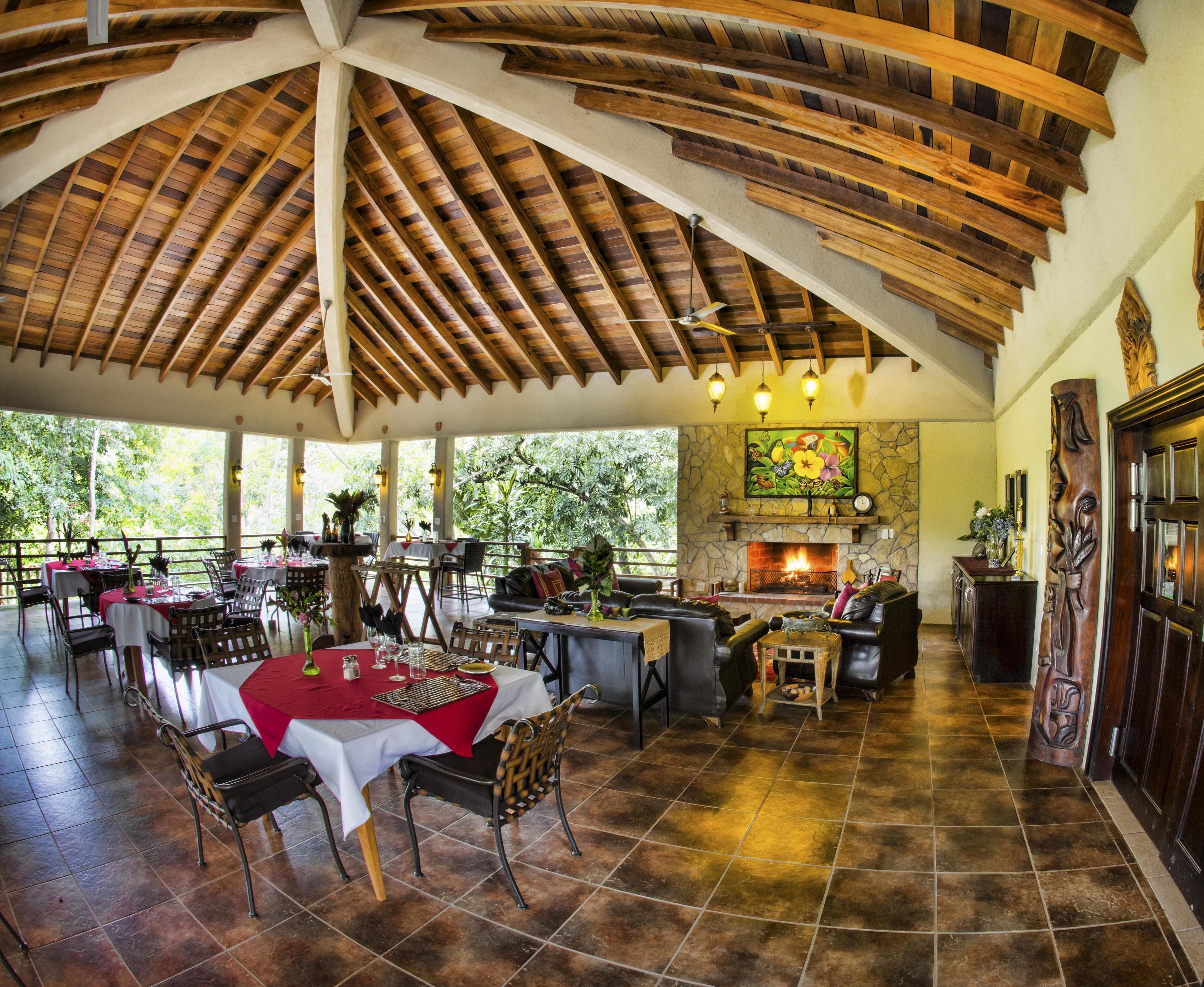 A spacious outdoor dining area with wooden beams, a stone fireplace, and a view of the surrounding greenery.