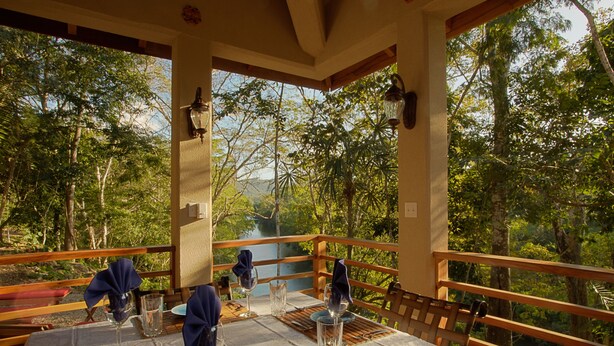 An outdoor dining area with a table set for four, surrounded by a wooden railing, overlooking a forested area with a river in the background. - Mystic River Resort (San Ignacio)