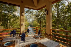 Un área de comedor al aire libre con una mesa preparada para cuatro personas, rodeada por una barandilla de madera, con vista a una zona boscosa que incluye un río al fondo.