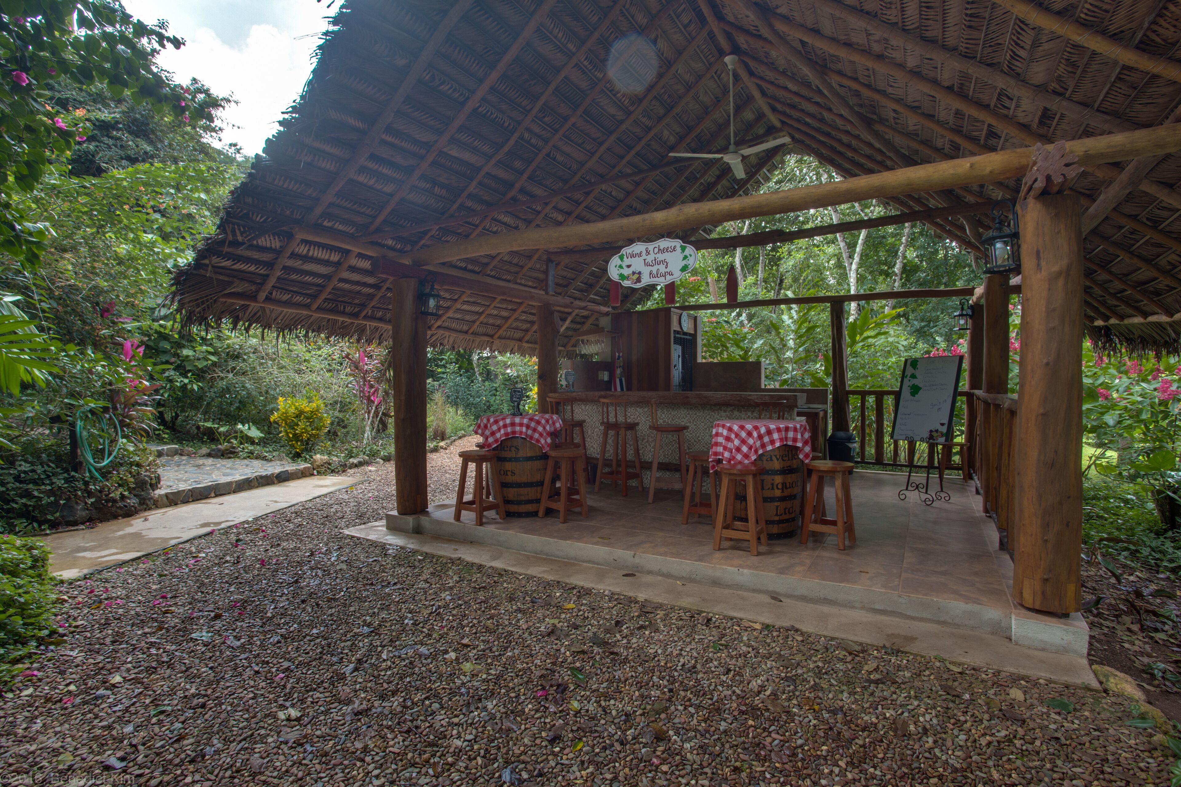 An outdoor café with a thatched roof, wooden tables, and chairs, surrounded by lush greenery.