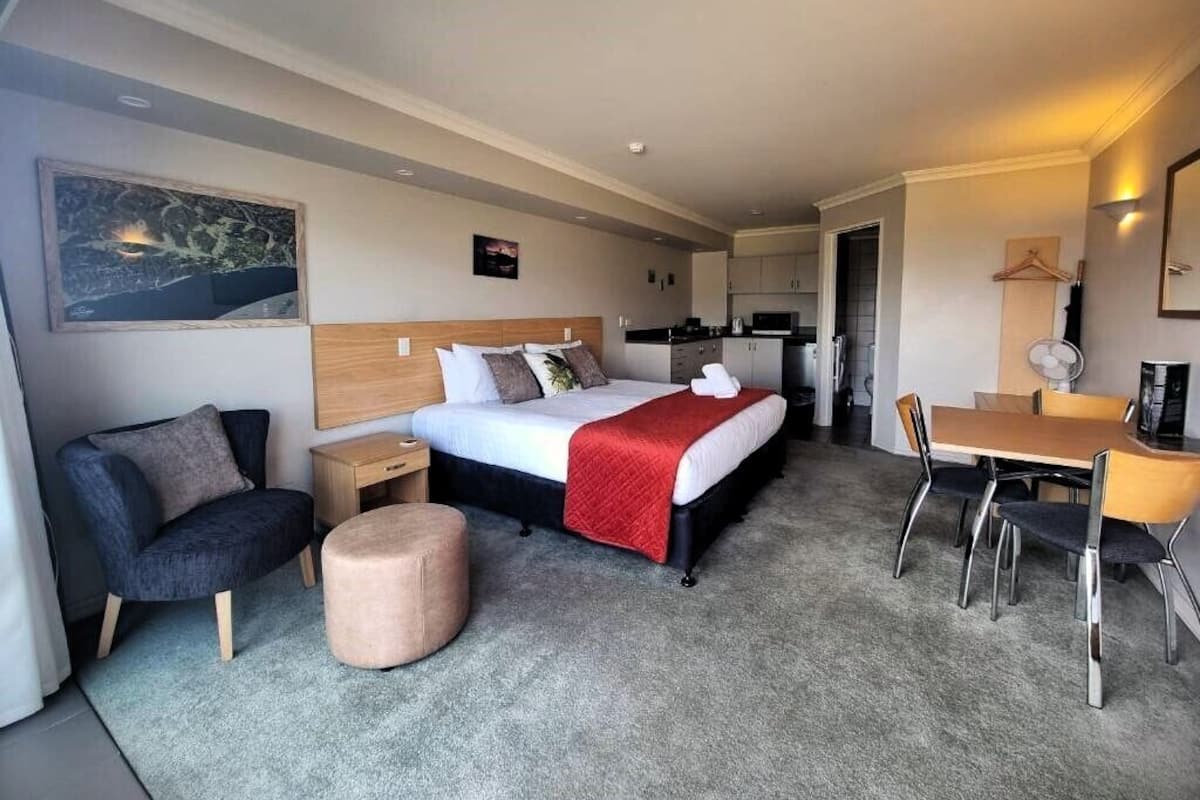 A hotel room with a bed, bedside table, chair, ottoman, and a dining area with a table and chairs.