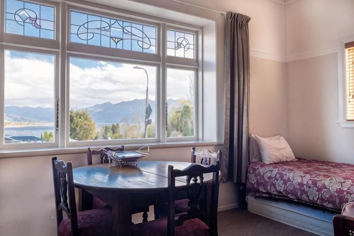 A room with a bed, a dining table with chairs, and a large window with a view of mountains and a lake.