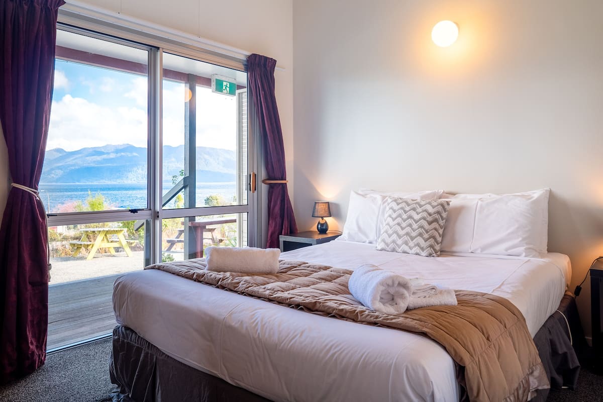 A bedroom with a large bed, a nightstand, a lamp, and a view of the outdoors.