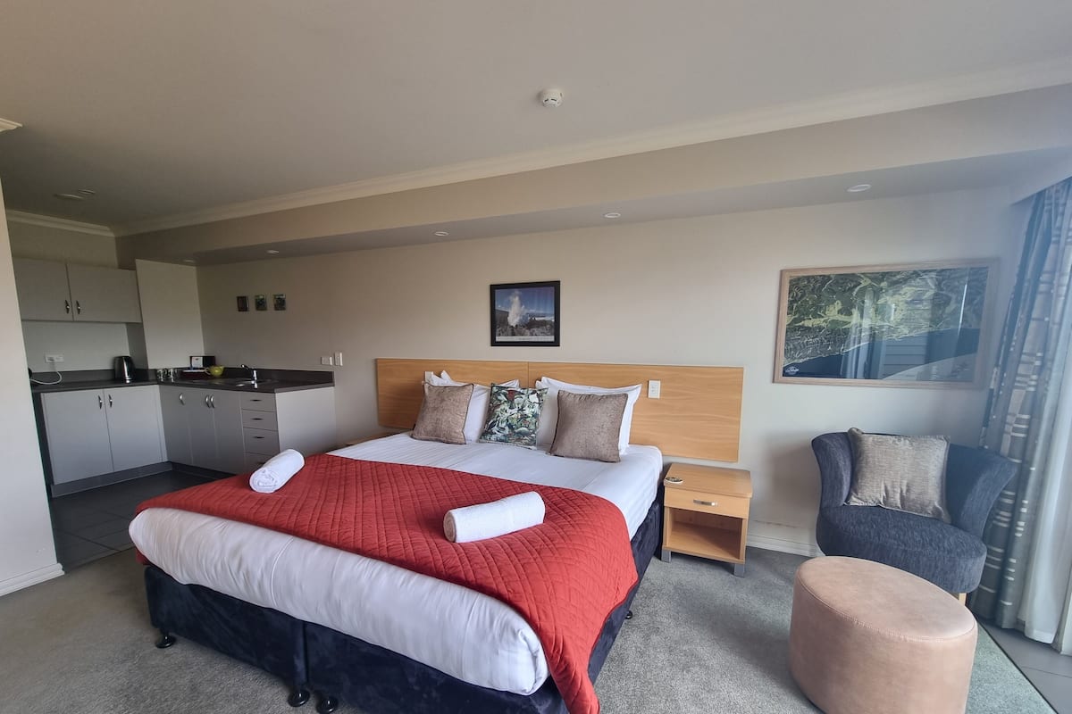 A modern hotel room with a large bed, a wooden headboard, a small desk, a chair, and a view of the outdoors.
