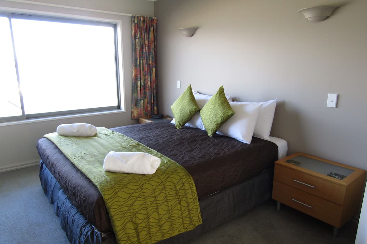 A neatly made bed with a green and brown bedspread, two green pillows, and a white towel. There is a small wooden nightstand with a drawer and a single light fixture on the wall.