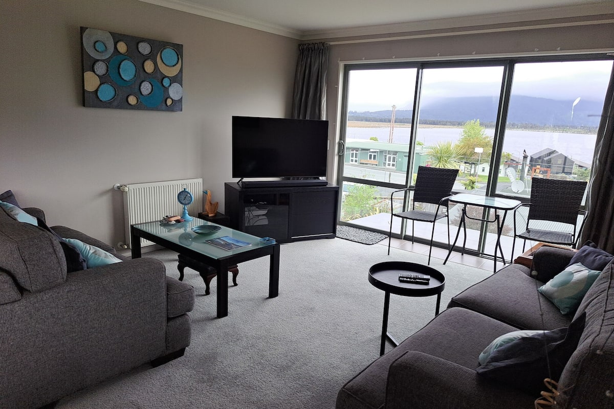 A living room with a sofa, a coffee table, a TV, and a view of a body of water through large windows.