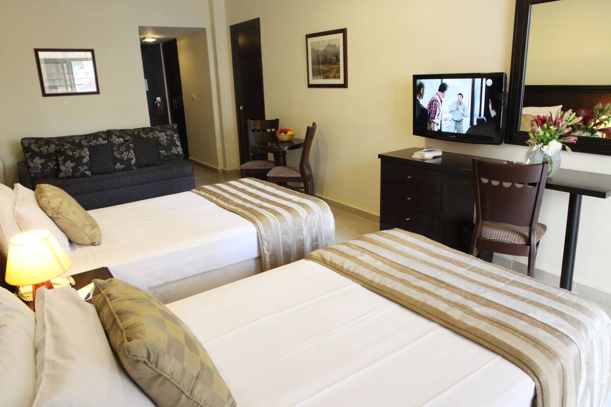 A hotel room with two beds, a desk with a television, a chair, and a small table with a fruit bowl.