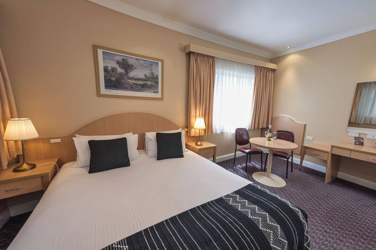 A hotel room with a large bed, two bedside tables with lamps, a desk, a chair, and a window with curtains.