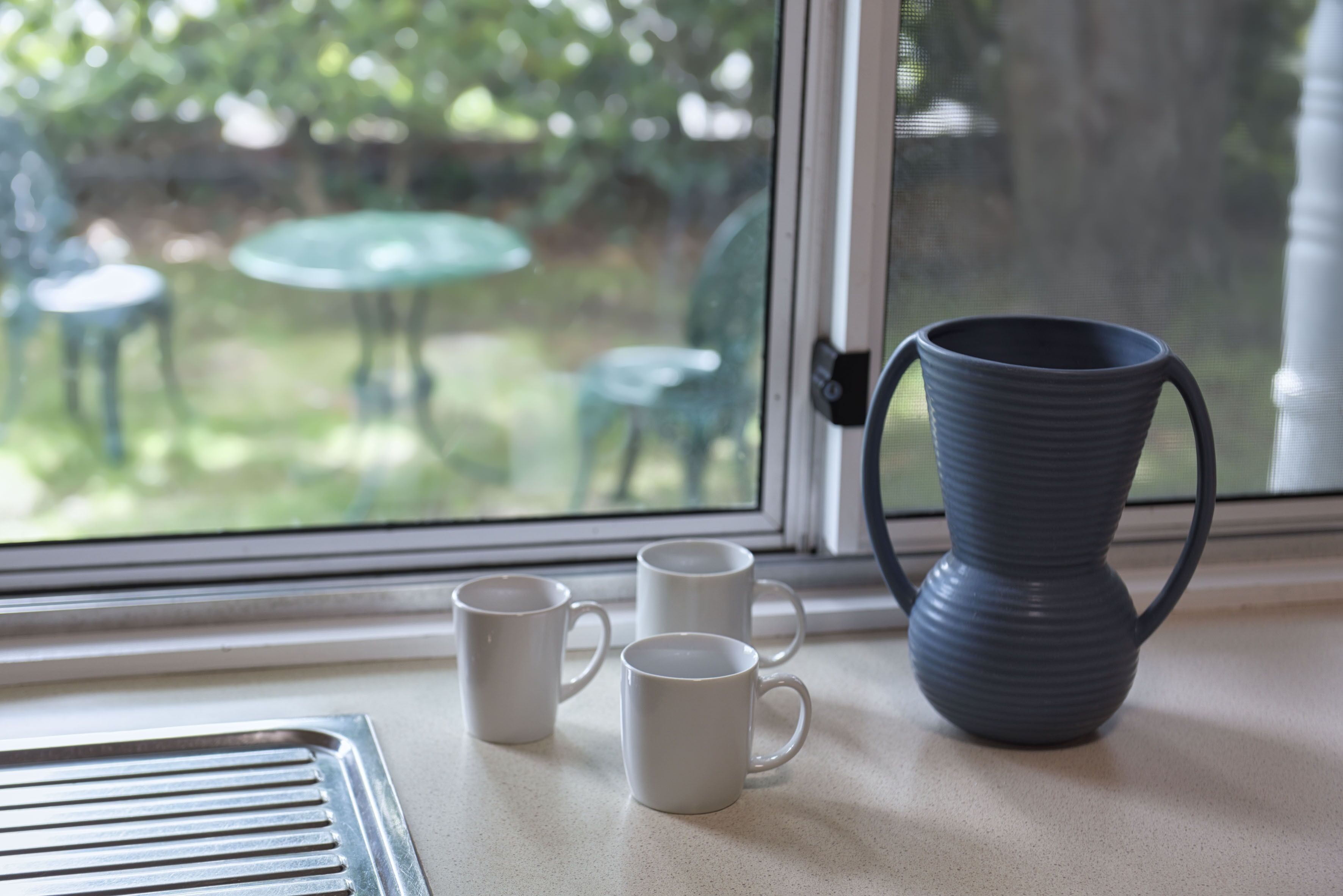 Tres tazas blancas sobre una encimera cerca de una ventana que ofrece vistas a muebles de exterior.