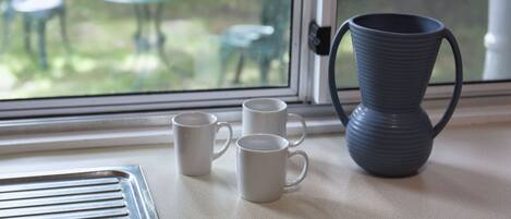 Tres tazas blancas sobre una encimera cerca de una ventana que ofrece vistas a muebles de exterior.