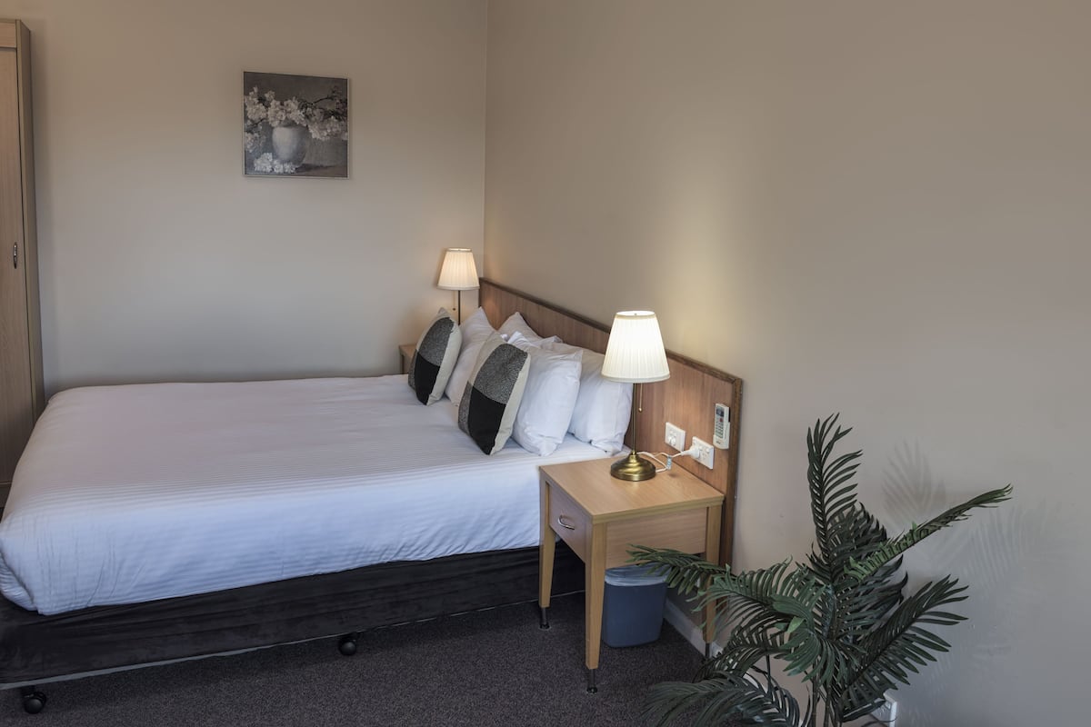 A neatly made bed with white linens, two bedside lamps, a small wooden nightstand, a trash bin, a potted plant, and a painting on the wall.