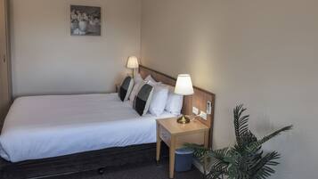 Un lit bien fait, avec du linge de lit blanc, deux lampes de chevet, une petite table de nuit en bois, une poubelle, une plante en pot et un tableau accroché au mur.