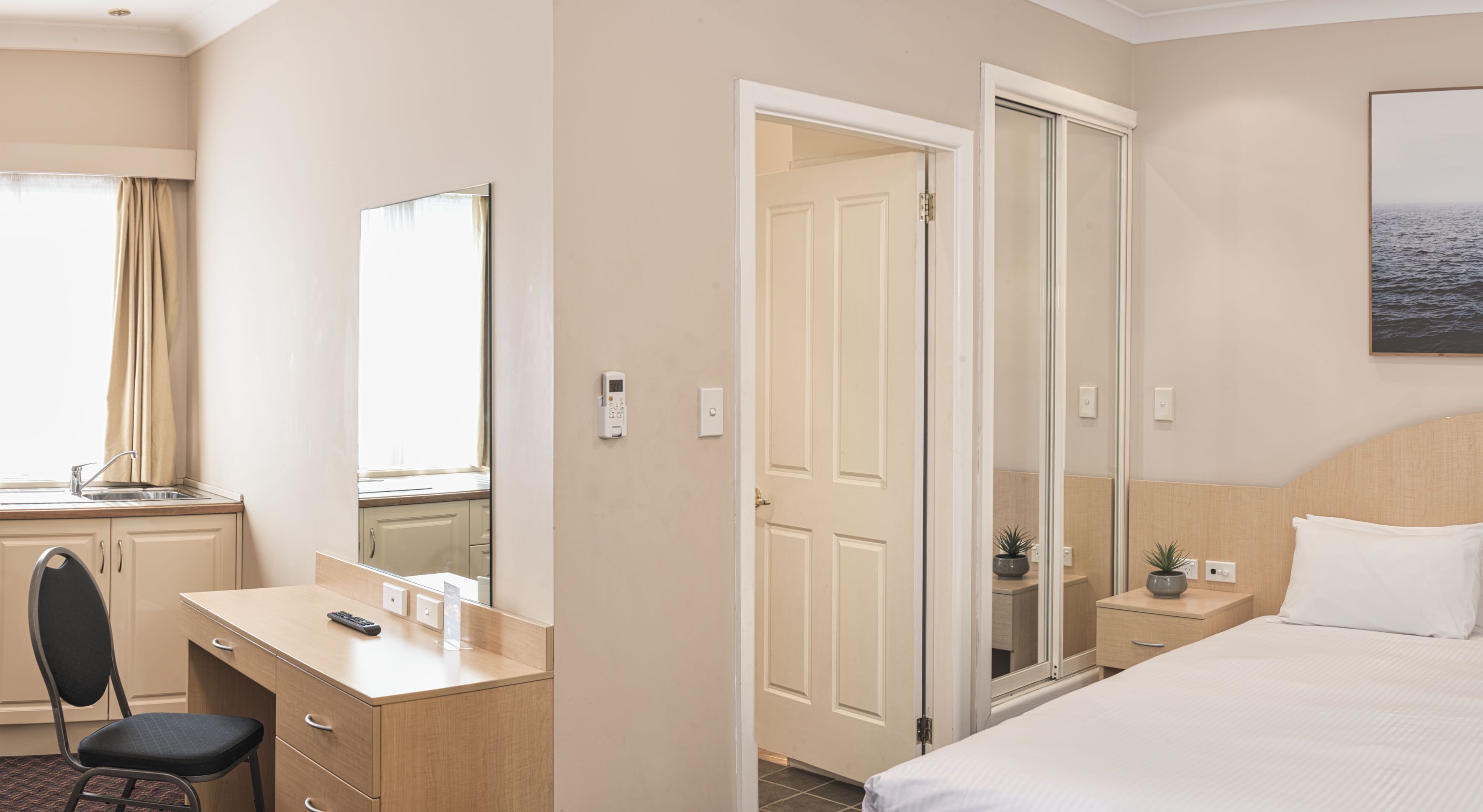 Habitación de hotel con cama, escritorio con silla, espejo, puerta y una pequeña planta en maceta.