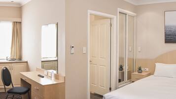 Habitación de hotel con cama, escritorio con silla, espejo, puerta y una pequeña planta en maceta.