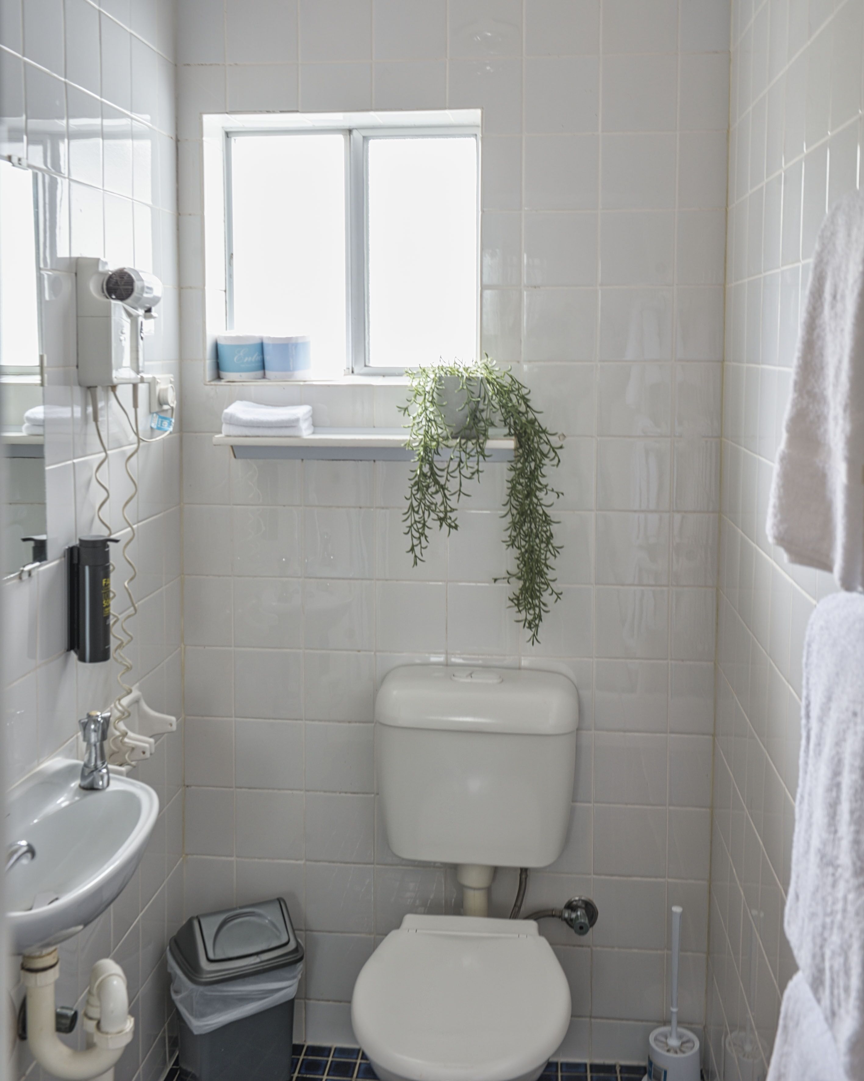 A small, modern bathroom with white tiles, a toilet, a sink, a window with a plant, and a towel hanging on the wall.
