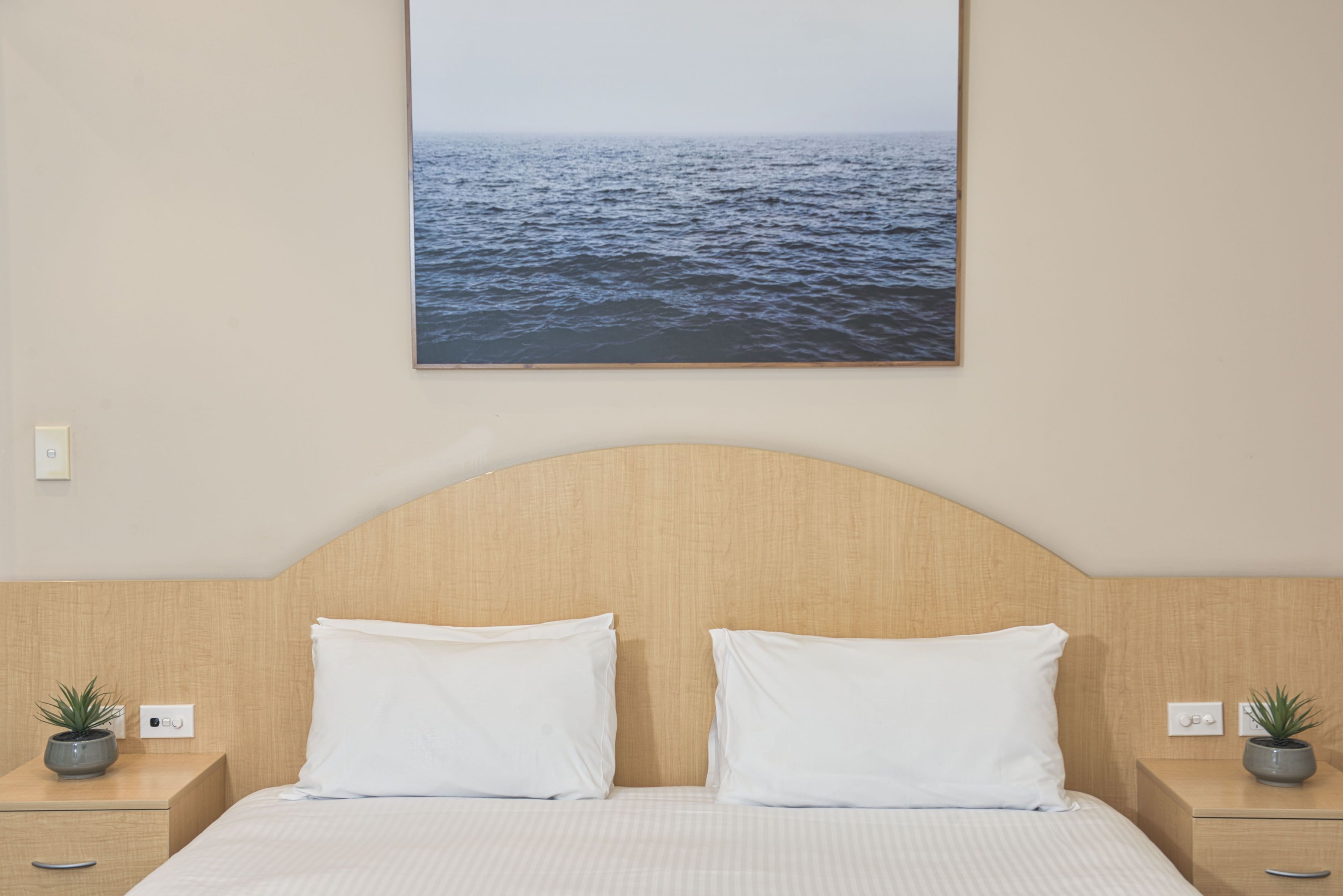 A hotel room with a large bed, two white pillows, a wooden headboard, and a wall-mounted picture of the sea.