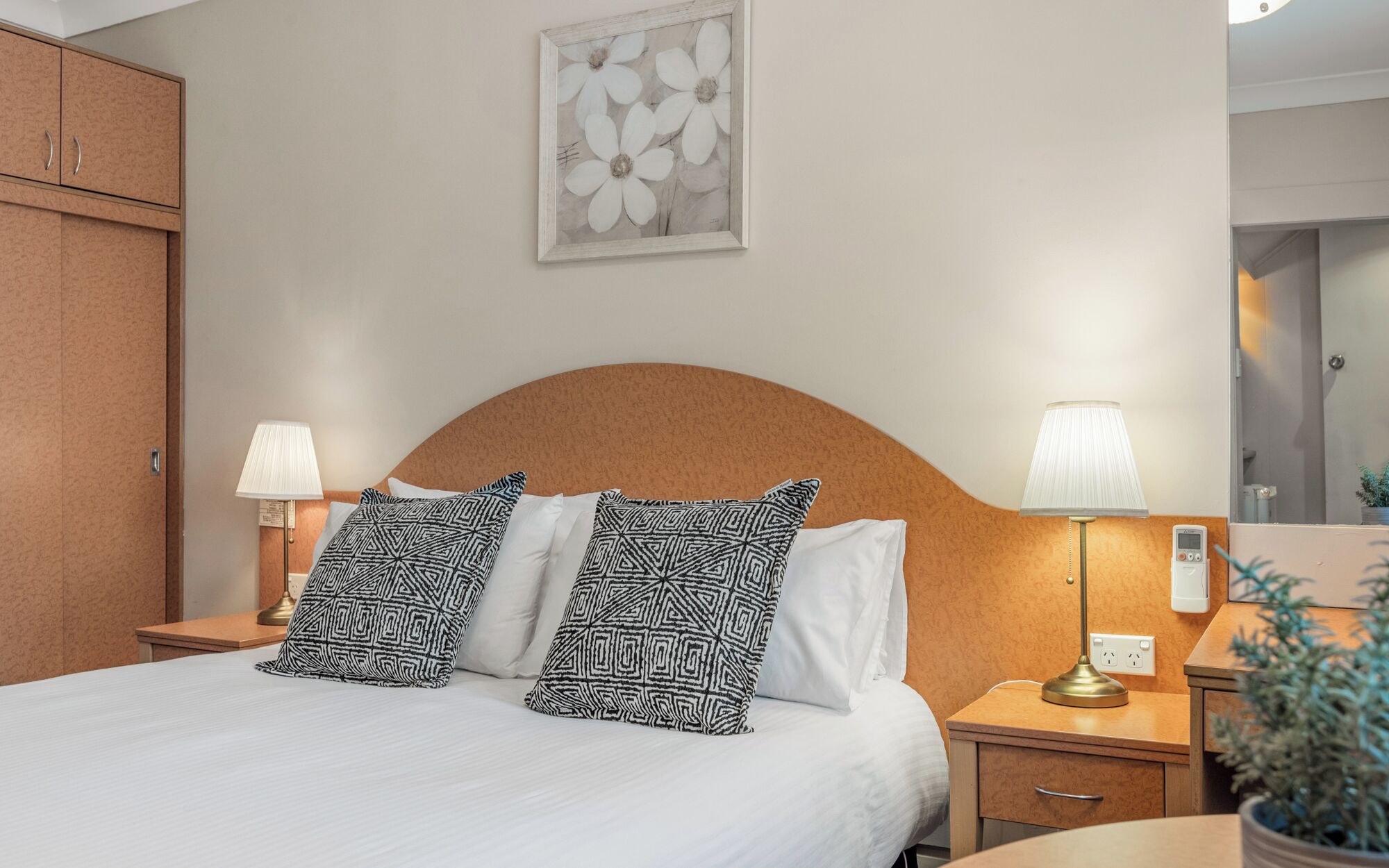 A hotel room with a bed, two bedside lamps, a nightstand, a potted plant, and a framed artwork on the wall.