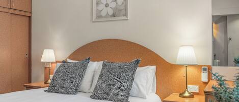 A hotel room with a bed, two bedside lamps, a nightstand, a potted plant, and a framed artwork on the wall.