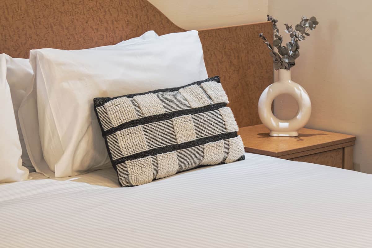 A neatly made bed with white linens and a black and white patterned pillow. A small white vase with a plant is on a wooden nightstand beside the bed.