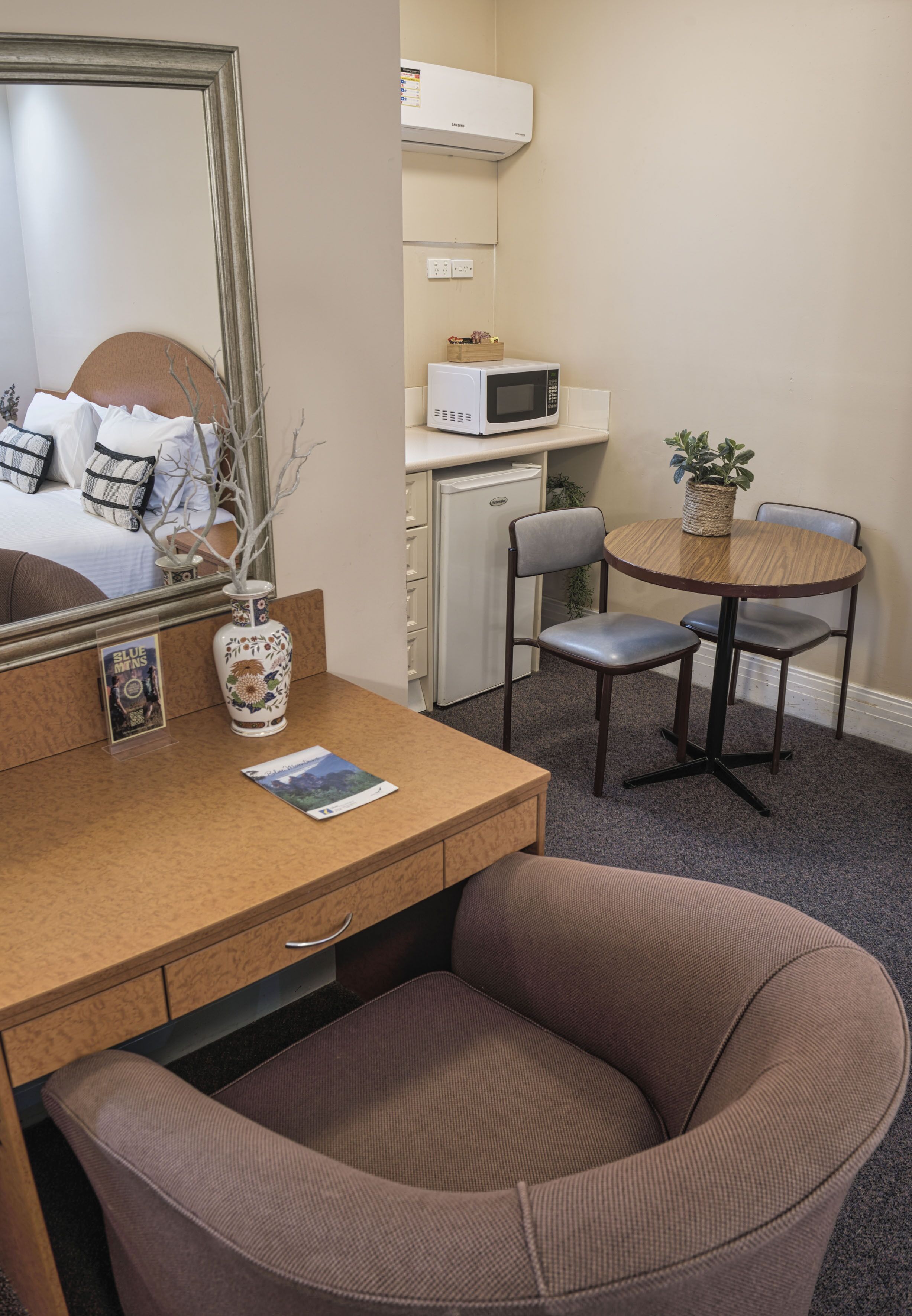 A hotel room with a bed, a desk with a mirror, a microwave, a small table with chairs, and a potted plant.