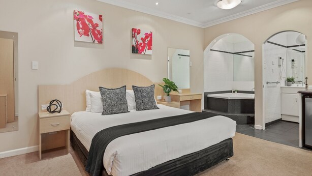A modern hotel room with a large bed, bedside tables, a desk, a vanity with a mirror, and a bathroom with a bathtub and shower. - Blue Mountains Heritage Motel (Katoomba)