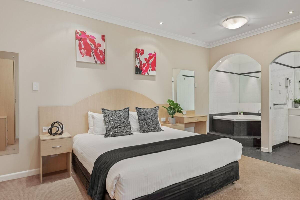 A modern hotel room with a large bed, bedside tables, a desk, a vanity with a mirror, and a bathroom with a bathtub and shower.