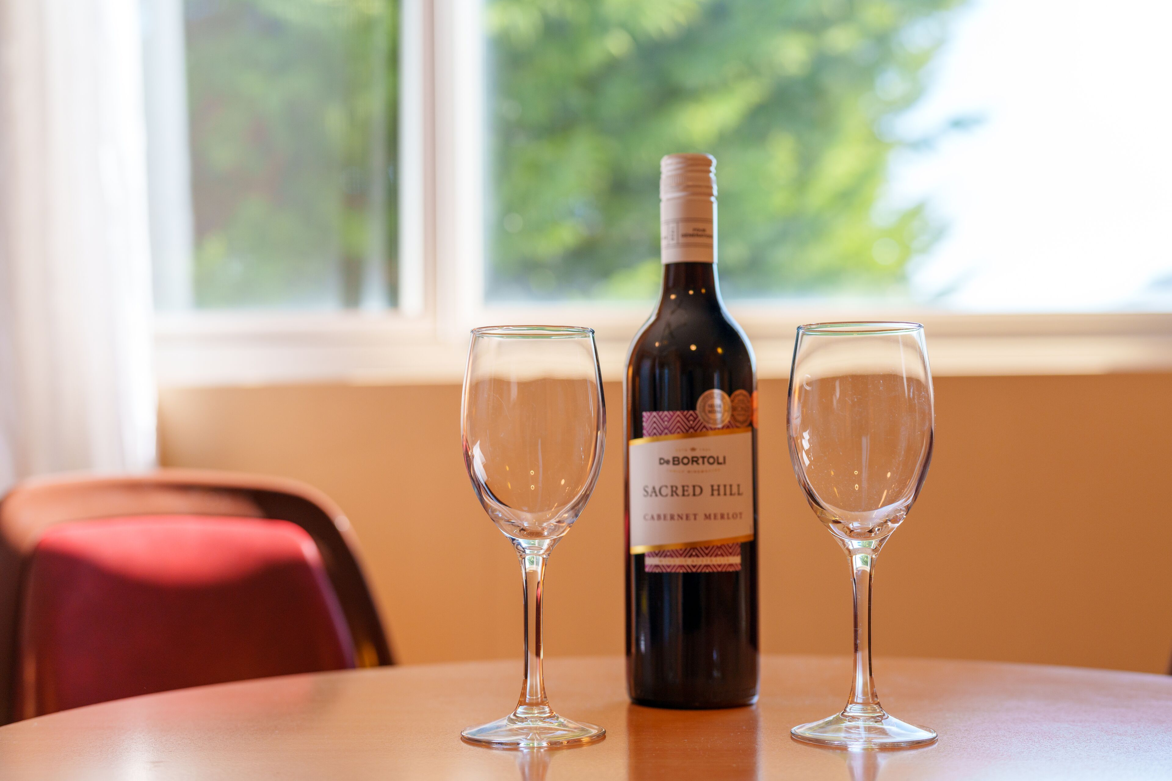 A bottle of wine and two glasses on a table by a window.