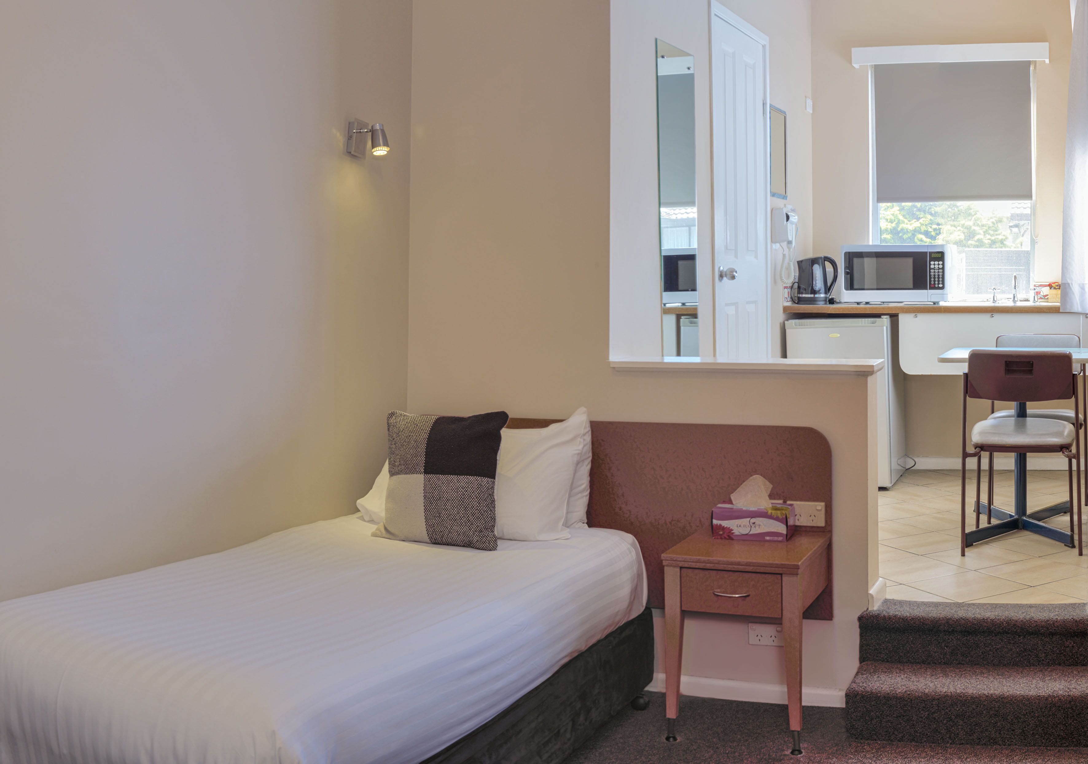 A small, compact room with a single bed, a bedside table with a lamp, a chair, and a kitchenette area with a microwave and a coffee maker.