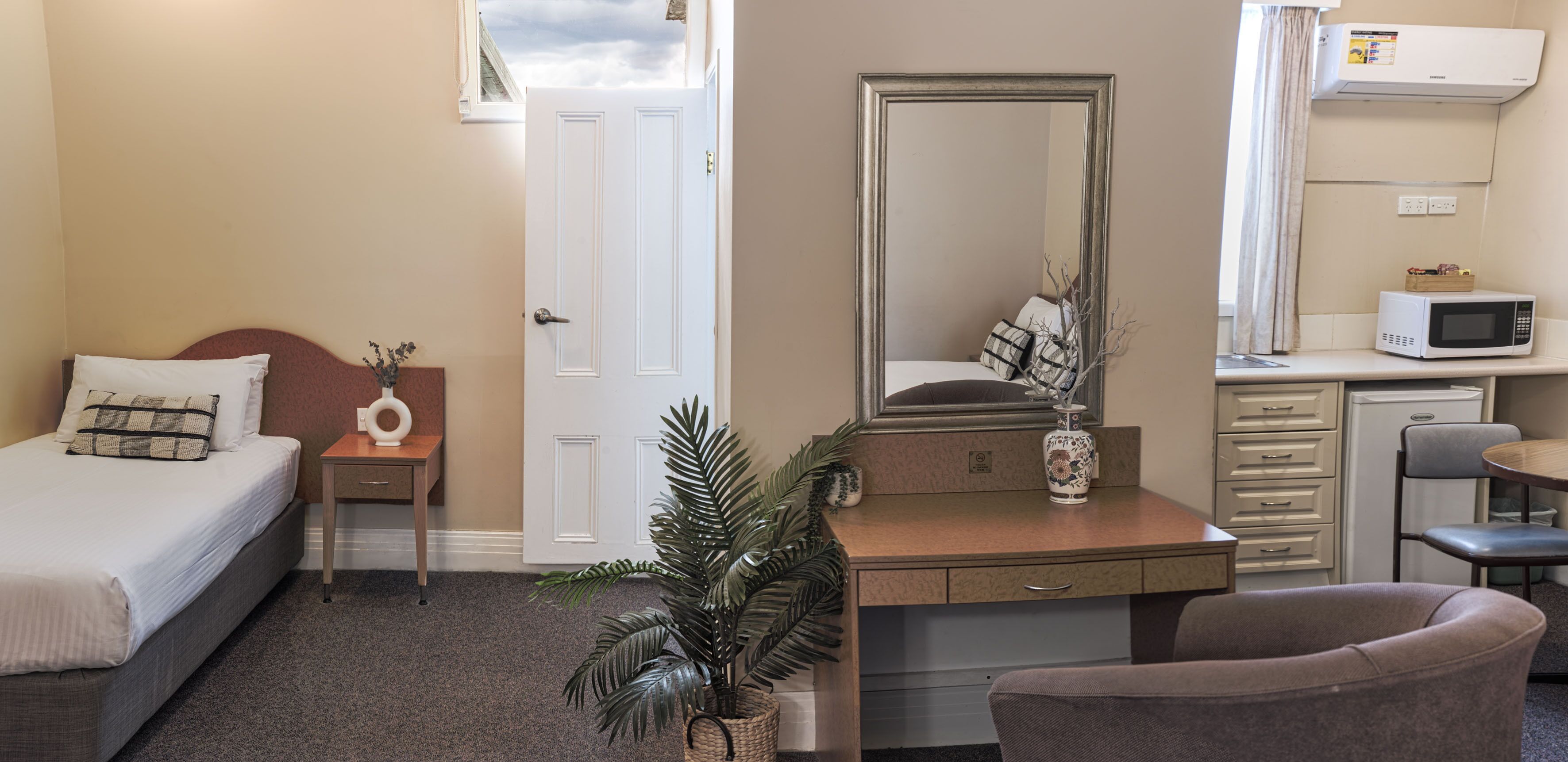 Une chambre d’hôtel équipée d’un lit, d’un bureau, d’une chaise, d’un miroir, d’une plante en pot, d’un four à micro-ondes et d’un petit réfrigérateur.