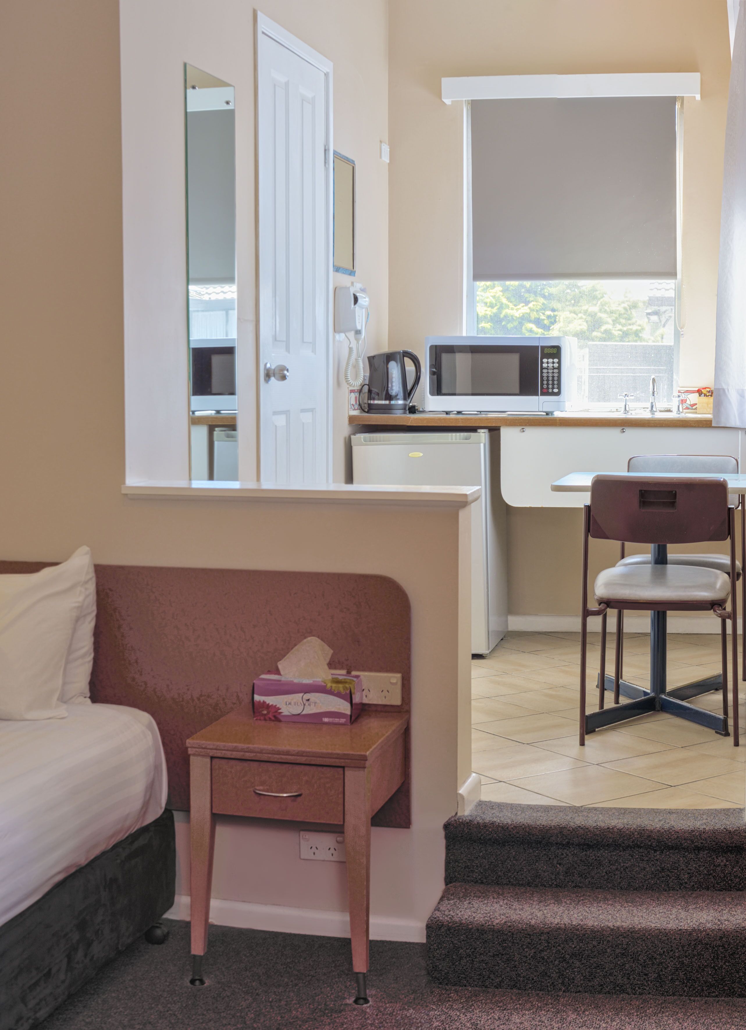 A hotel room with a bed, a desk with a microwave, a chair, a small table with a tissue box, and a staircase.