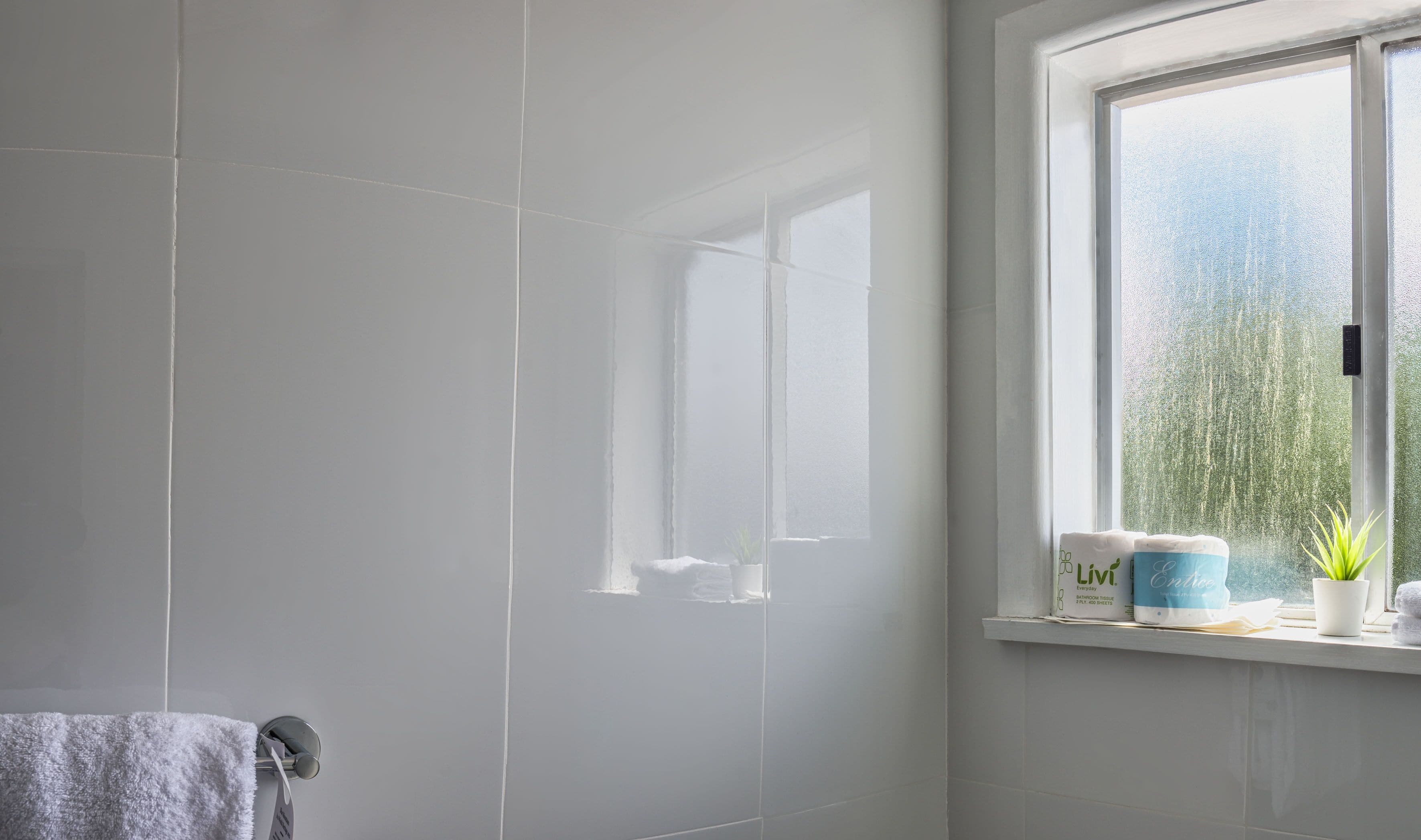 A bathroom with white tiles, a window with a view of greenery, and a shelf with toiletries.