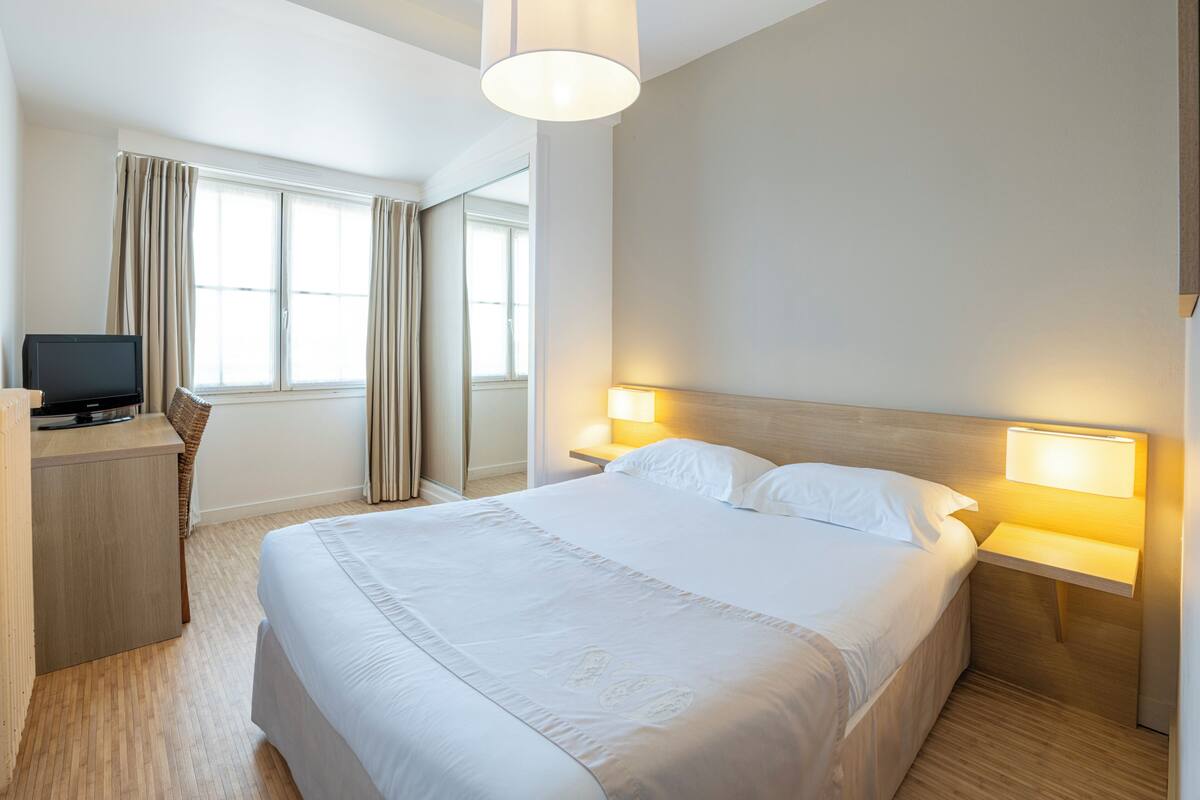A hotel room with a bed, bedside lamps, a desk with a computer, and a window with curtains.